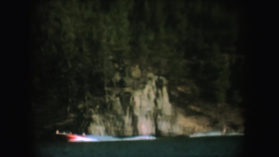 1954 South Dakota 8mm frame showing a small boat crossing shaded lake water near rocks and dense trees.
