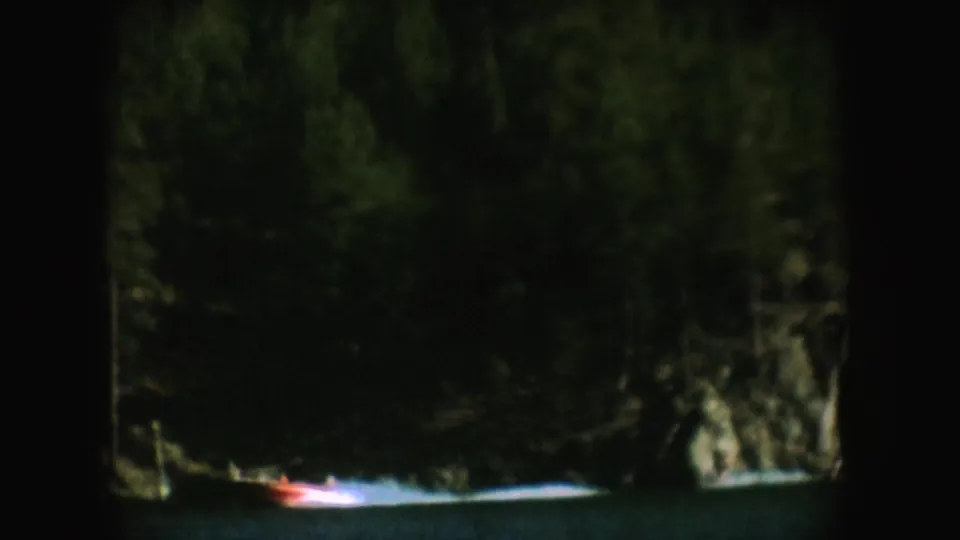 Small motorboat travels across the lake near a shadowed tree line and rocky bank in South Dakota home-movie footage.