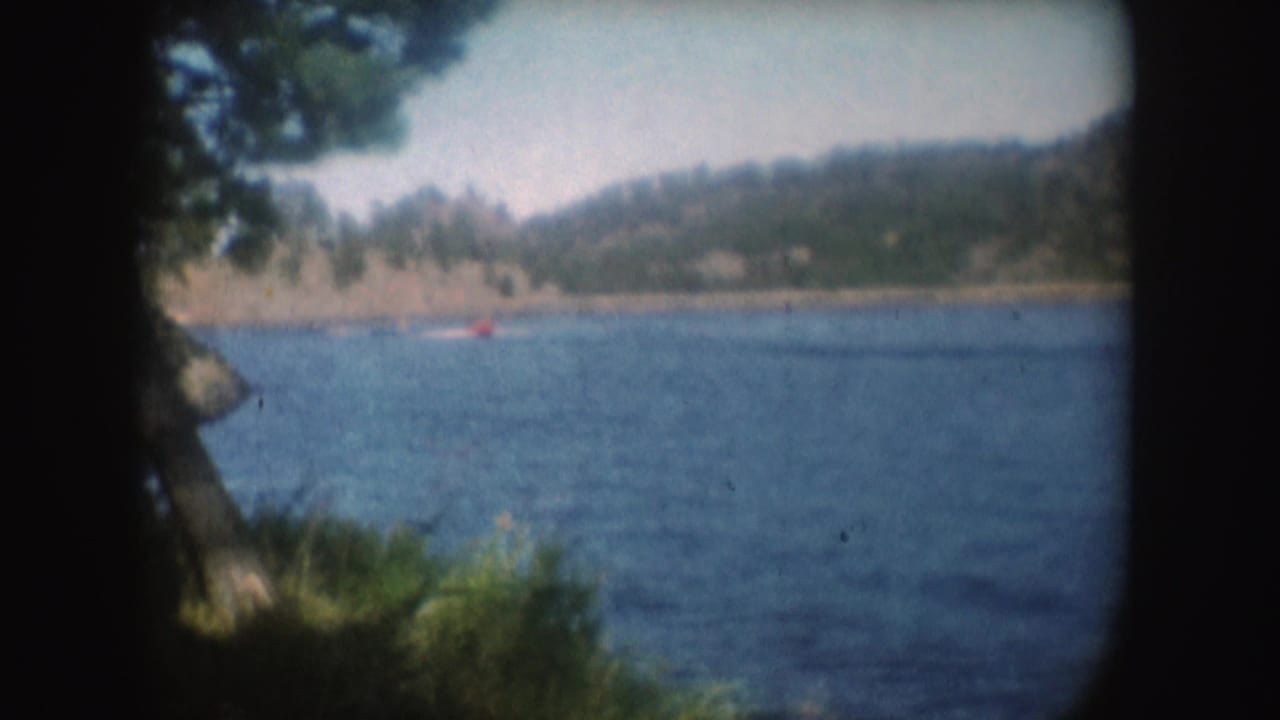 The boat appears farther across the South Dakota lake, with pale wake, wooded shoreline, and soft 8mm color visible.