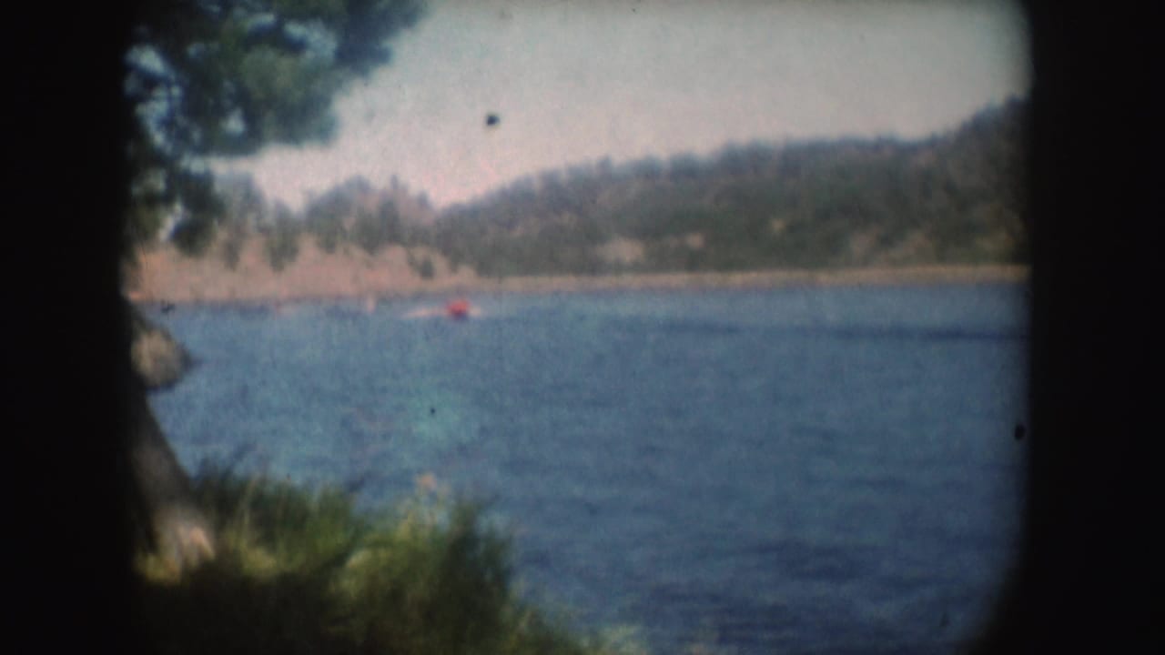 A distant red boat continues across the lake in this 1954 South Dakota home-movie view, framed by branches and soft foreground brush.