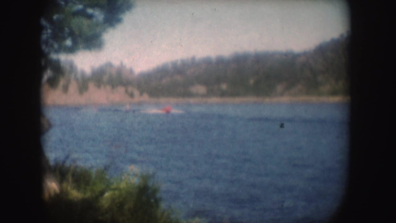 Late-frame 8mm view of the red boat and wake moving across a South Dakota lake, with trees and hills softened by film grain.