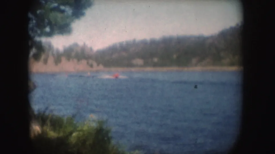 Late-frame 8mm view of the red boat and wake moving across a South Dakota lake, with trees and hills softened by film grain.