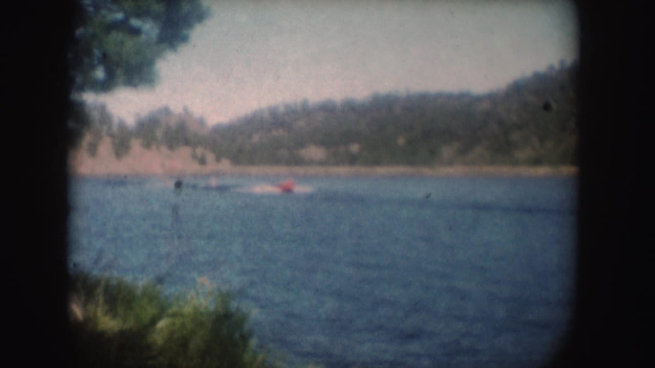 Small red boat moving across a lake with wooded hills beyond.