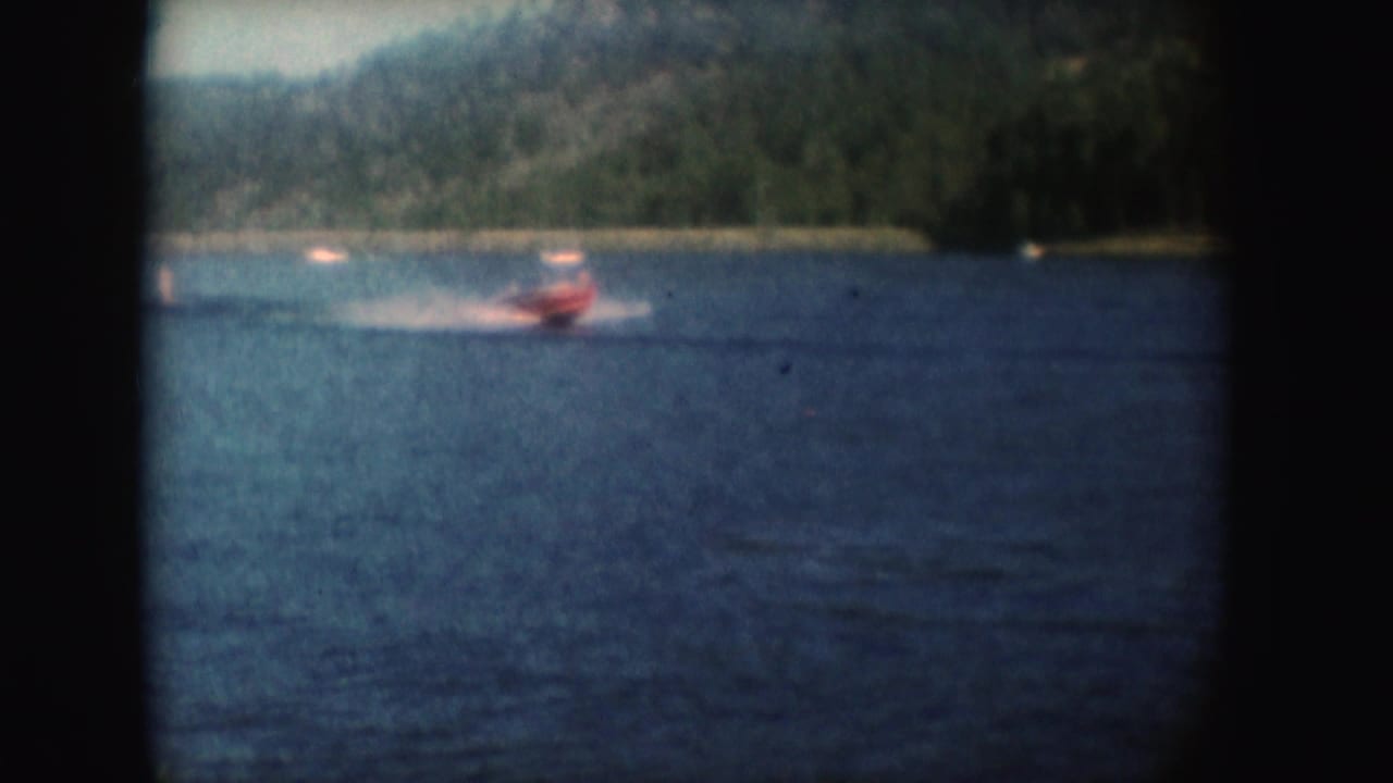 Red motorboat speeds left to right across a South Dakota lake, leaving a white wake below wooded hills.