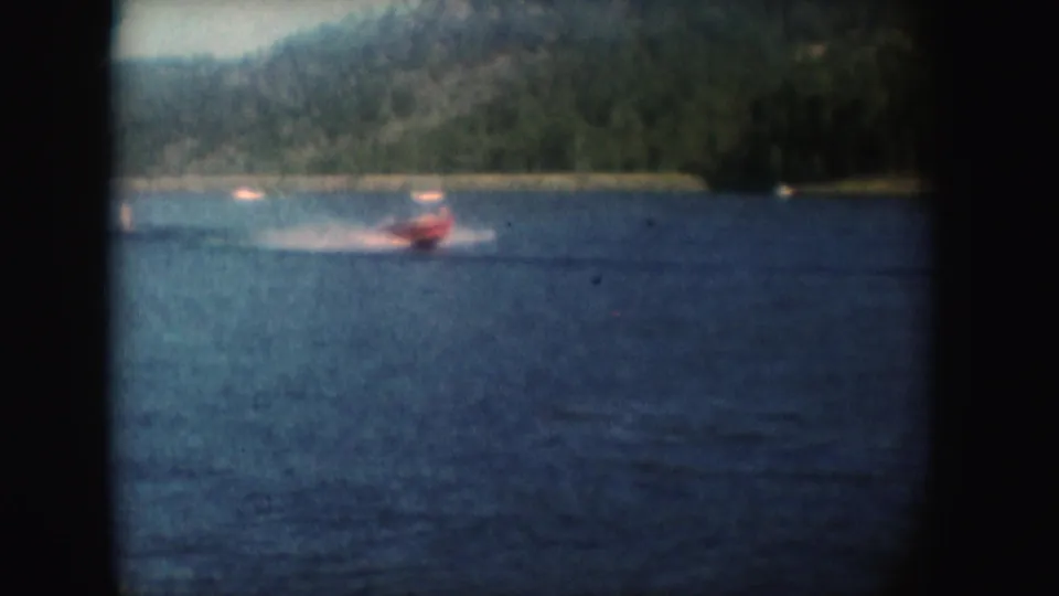 Red motorboat speeds left to right across a South Dakota lake, leaving a white wake below wooded hills.