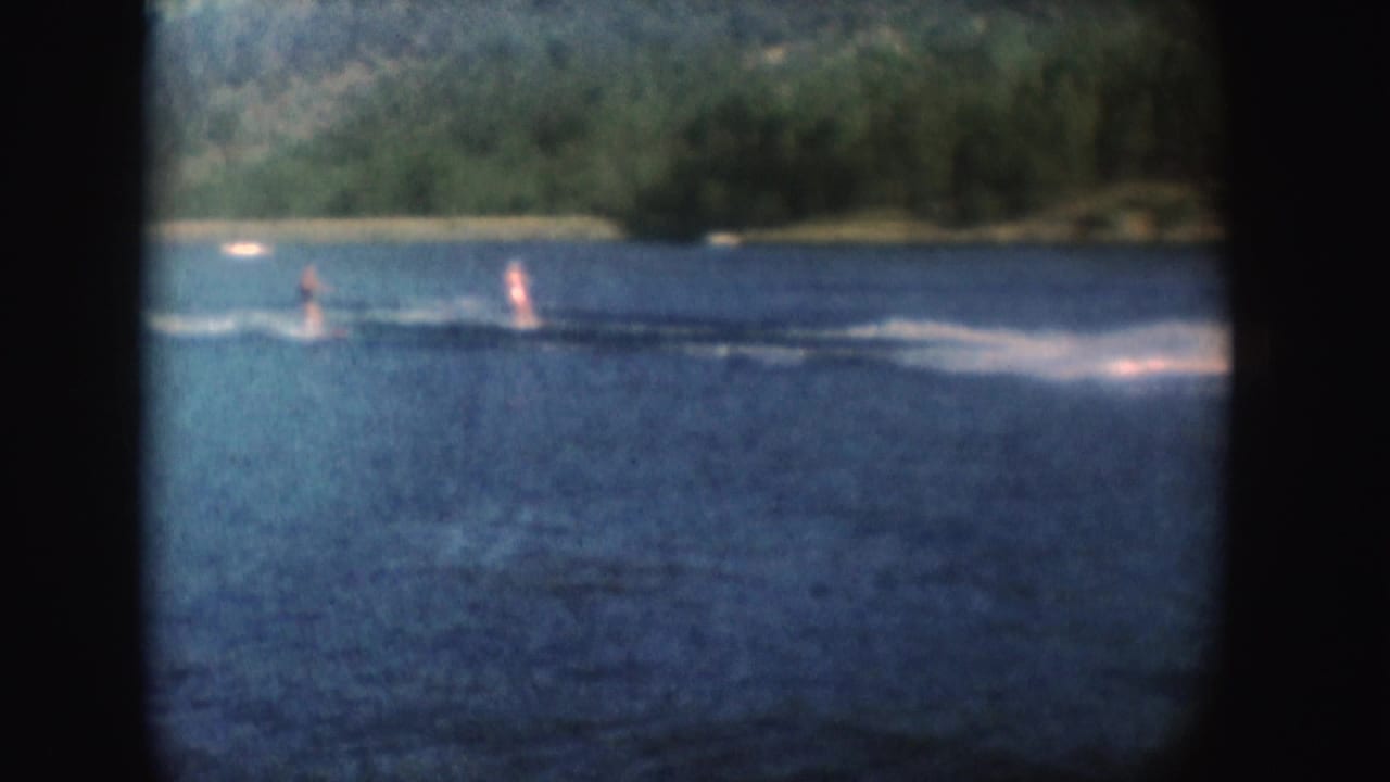 Two distant people water-ski across a South Dakota lake, with the towing boat mostly out of frame and forested shore behind.