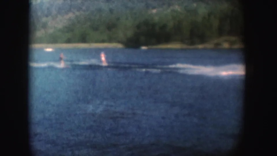 Two distant people water-ski across a South Dakota lake, with the towing boat mostly out of frame and forested shore behind.