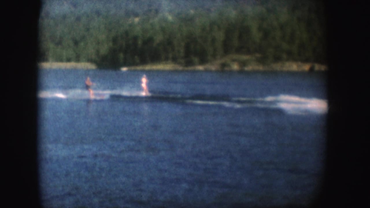 Two distant water skiers move across a blue lake near a tree-lined shore.