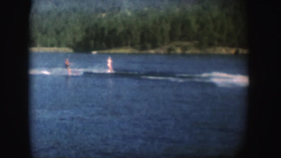 Two people water ski across a South Dakota lake in 1954, seen from a distant 8mm home-movie viewpoint with wooded shore behind them.