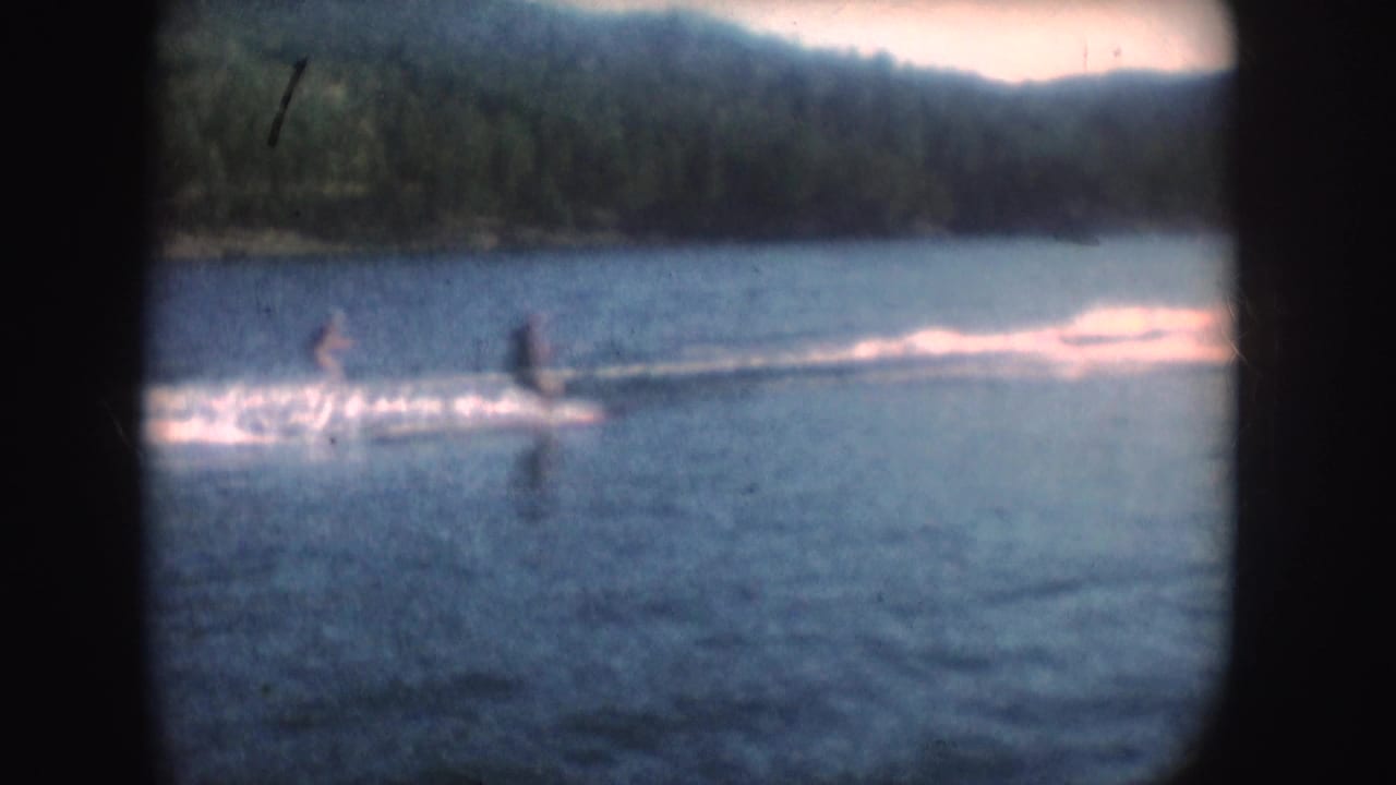 Distant water skiers continue across the lake in 1954 South Dakota footage, with bright wake lines and forested shore in the background.