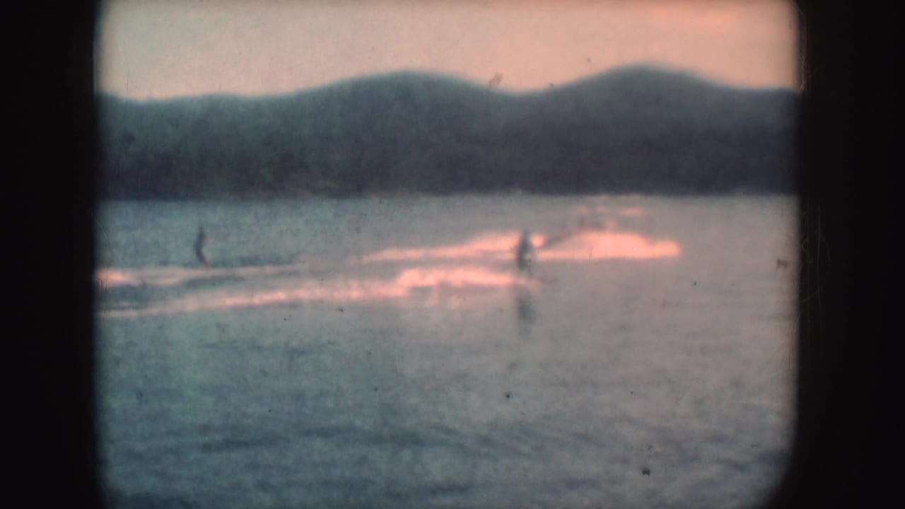 A skier crosses the lake in distant 1954 South Dakota home-movie footage, with another small figure, wake trails, hills, and faded color visible.