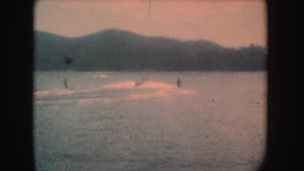 The water skiers appear farther away on the South Dakota lake, with fading 8mm color, soft hills, and long pale wake trails across the water.