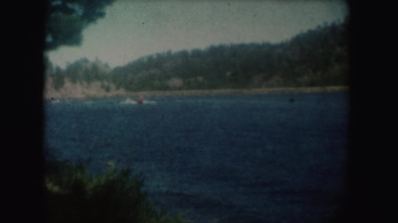 1954 South Dakota lake scene with small distant boats on blue water, a wooded hillside, shoreline, and vignetting from the 8mm frame.