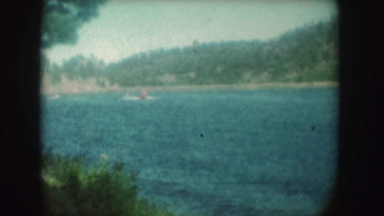 1954 South Dakota 8mm view across a lake toward wooded hills, with a small boat in the distance and soft foreground foliage.