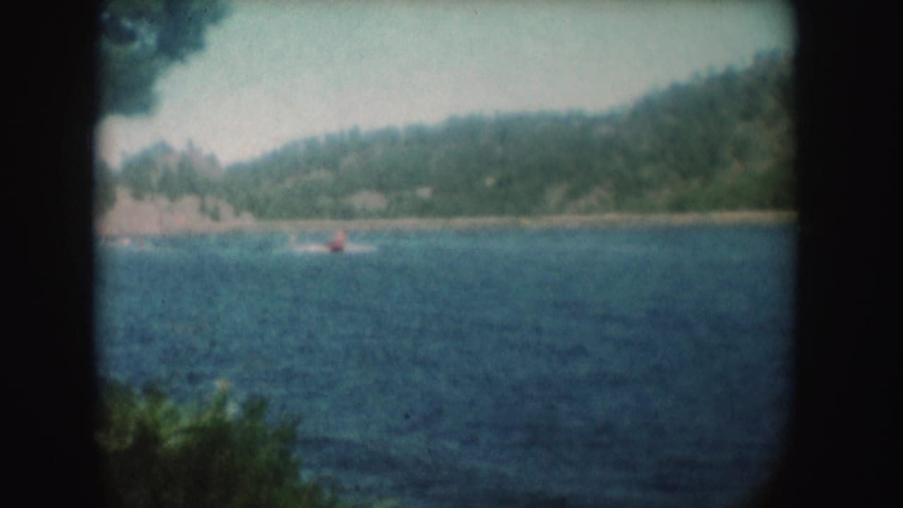1954 South Dakota lake footage showing a small red boat far from shore, wooded hills, rippled blue water, and soft 8mm vignetting.