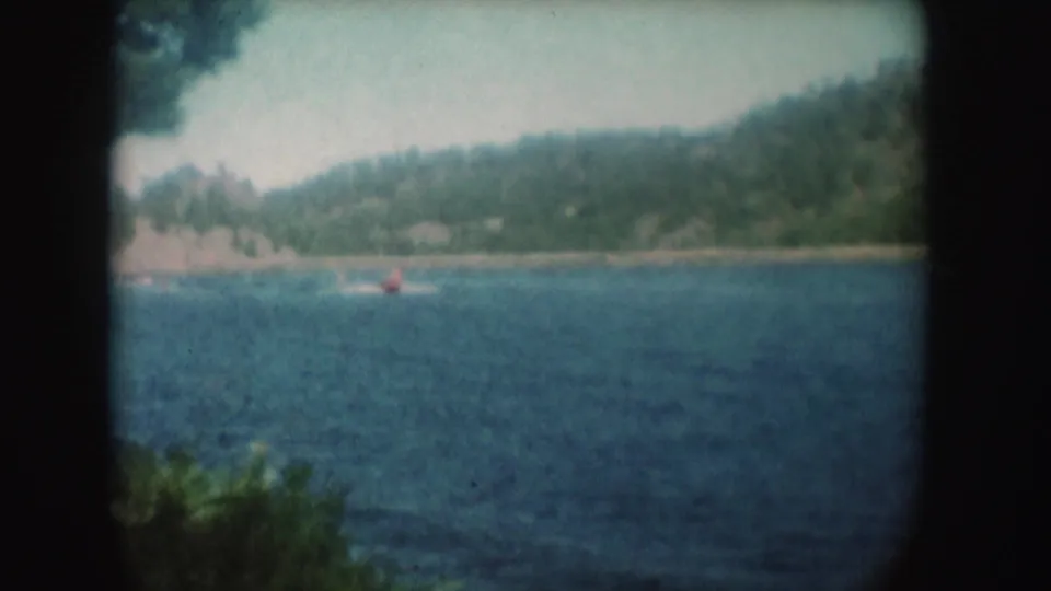 1954 South Dakota lake footage showing a small red boat far from shore, wooded hills, rippled blue water, and soft 8mm vignetting.