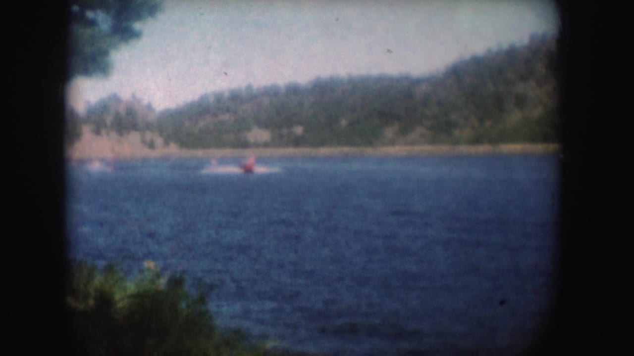 1954 South Dakota 8mm frame showing a small red speedboat far across a blue lake with wooded hills beyond.