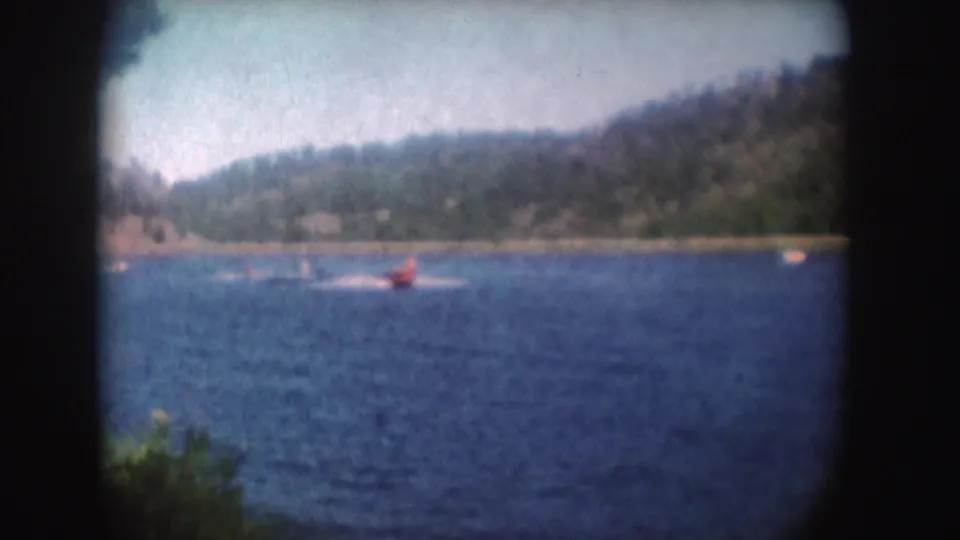 Distant 1954 South Dakota 8mm view of a red speedboat cutting across blue lake water near wooded hills.