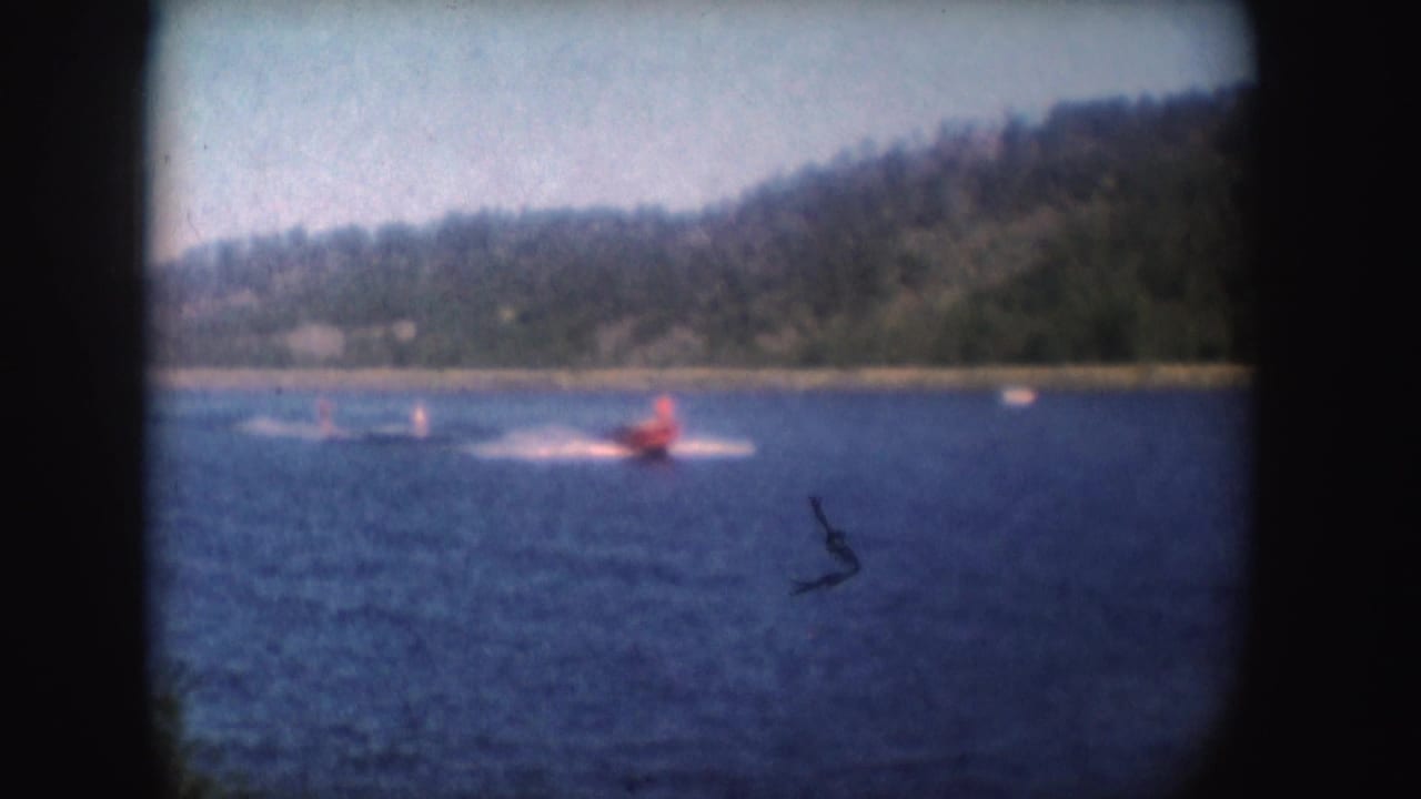 Red speedboat pulls two distant water skiers across a lake with wooded hills behind.