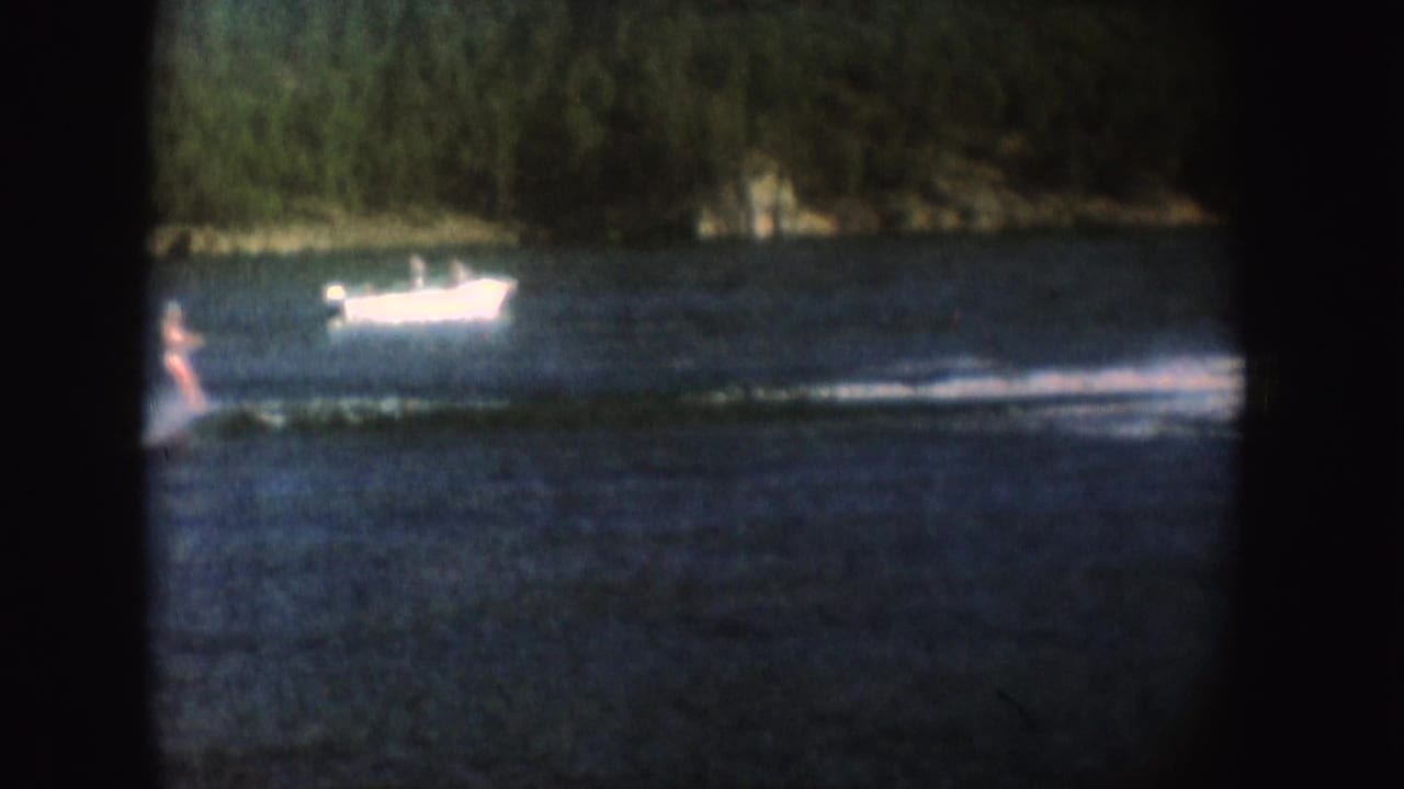 1954 South Dakota 8mm frame showing a motorboat pulling a water skier across a lake near a wooded shore.