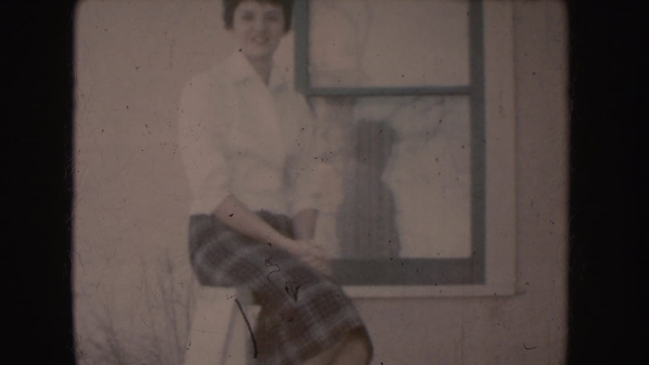 Young woman in a blouse and plaid skirt sits on a stool beside a window.
