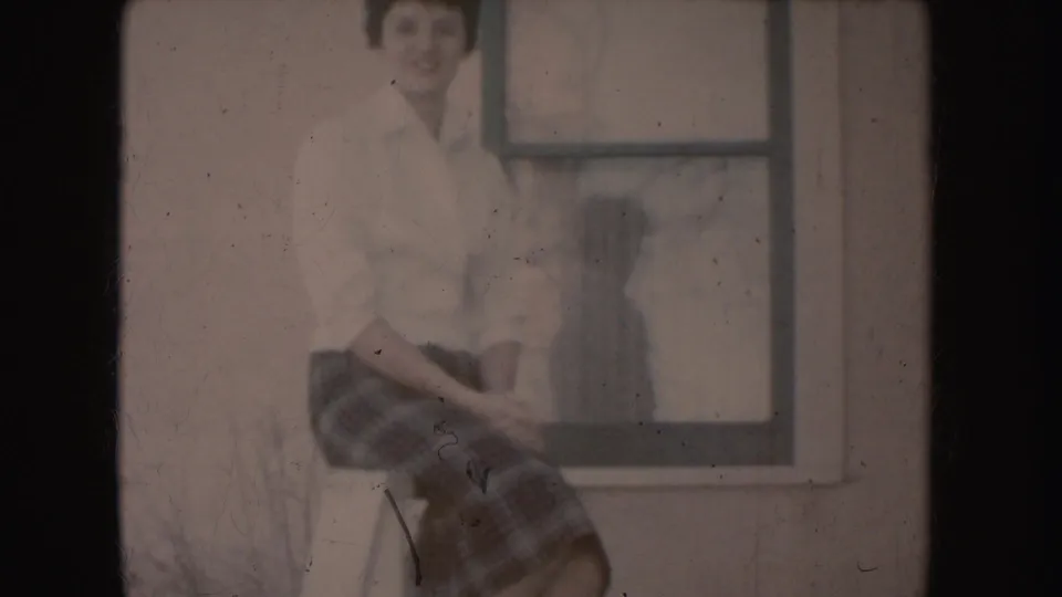Young woman in 1950s attire sits outside a South Dakota building, smiling faintly beside a window in faded 8mm color.