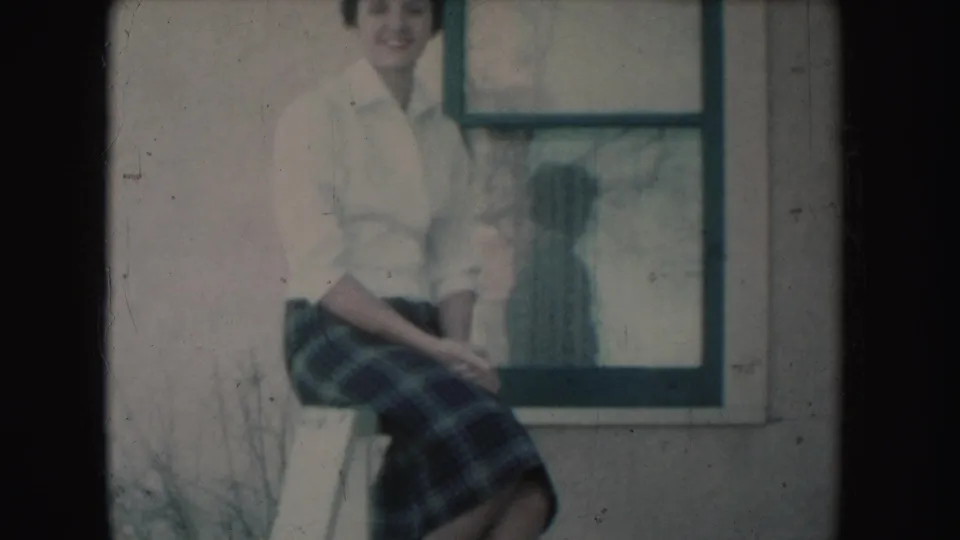 In this 1954 South Dakota home-movie frame, a seated young woman smiles outside beside a window with a dark reflection or shadow.