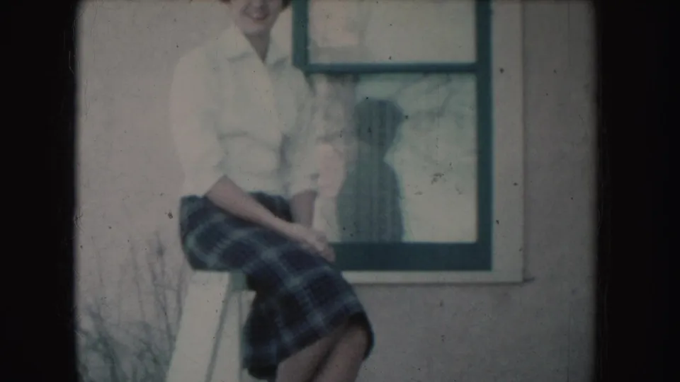 A 1954 South Dakota 8mm frame shows the seated young woman partly cropped, with plaid skirt, white blouse, window, and wall visible.