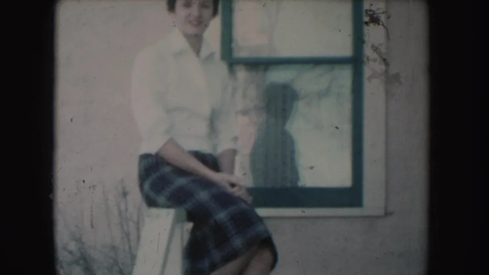 The young woman remains seated outside a South Dakota building in 1954, wearing a white blouse and plaid skirt beside the window.