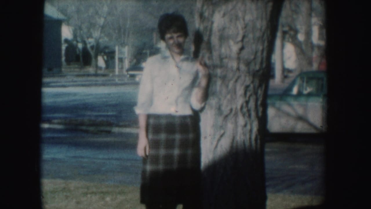 Adult woman in 1954 South Dakota home-movie footage stands beside a tree on a quiet residential street.
