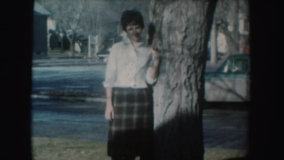 Adult woman in 1954 South Dakota home-movie footage stands beside a tree on a quiet residential street.