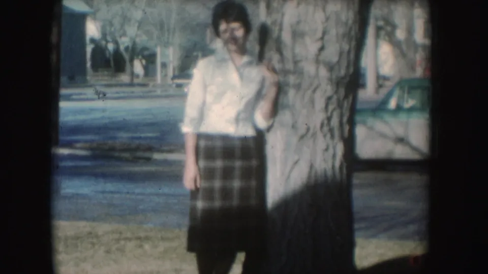 Soft 8mm frame of the woman beside the tree, her clothing and the nearby street softened by grain and fading.