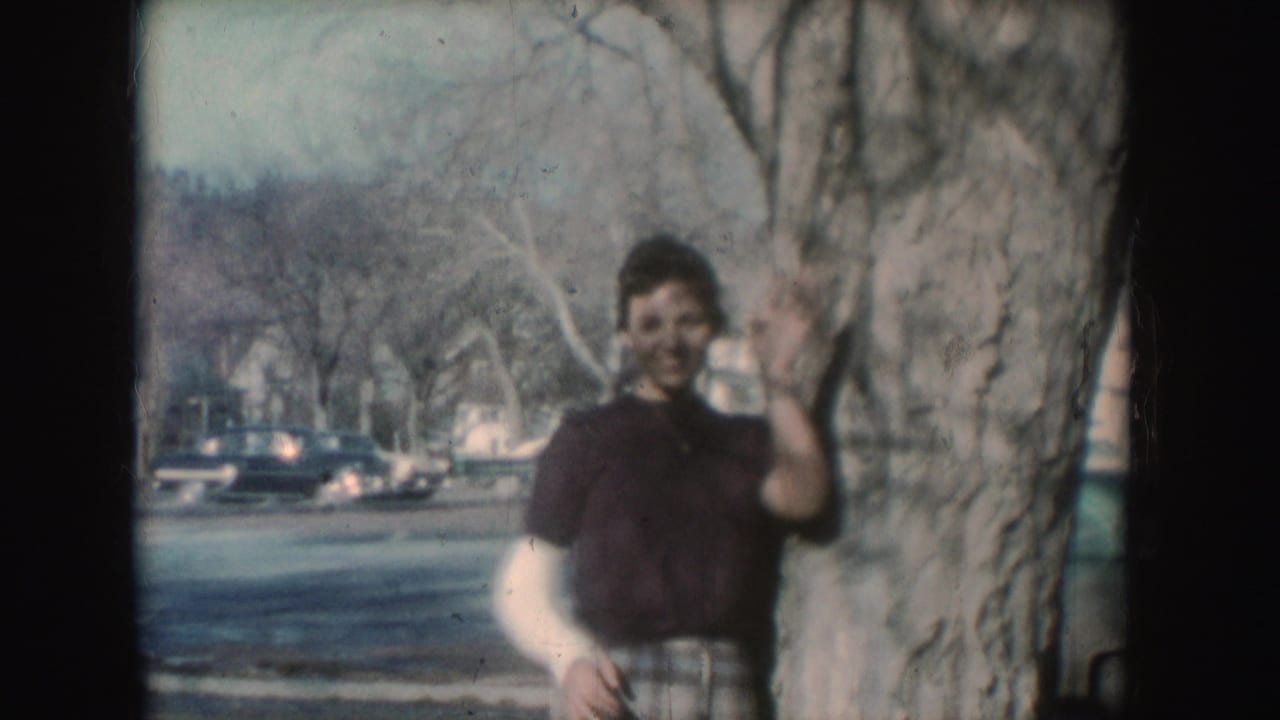 Young woman with a white arm cast waves beside a large tree on a street.