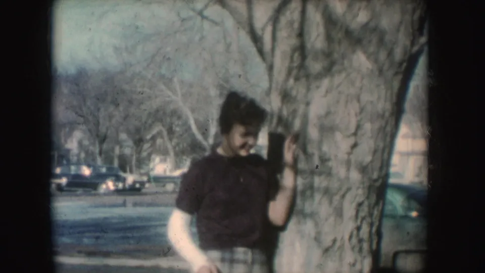 The woman in a white cast leans against a tree in a South Dakota street scene, with parked cars and bare branches behind her.