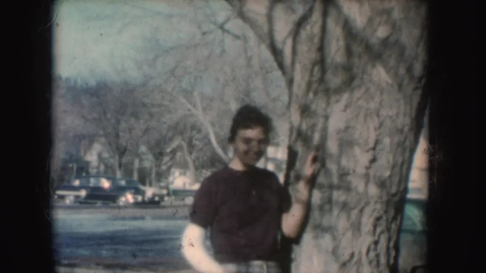 A young woman in 1954 South Dakota footage smiles near a large tree, wearing plaid pants and a dark top while her cast is visible.