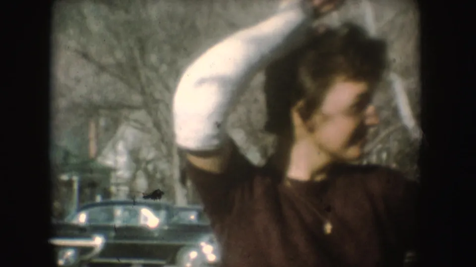 The camera moves closer as the woman raises her cast near her head, with mid-century cars and winter-bare trees in the background.