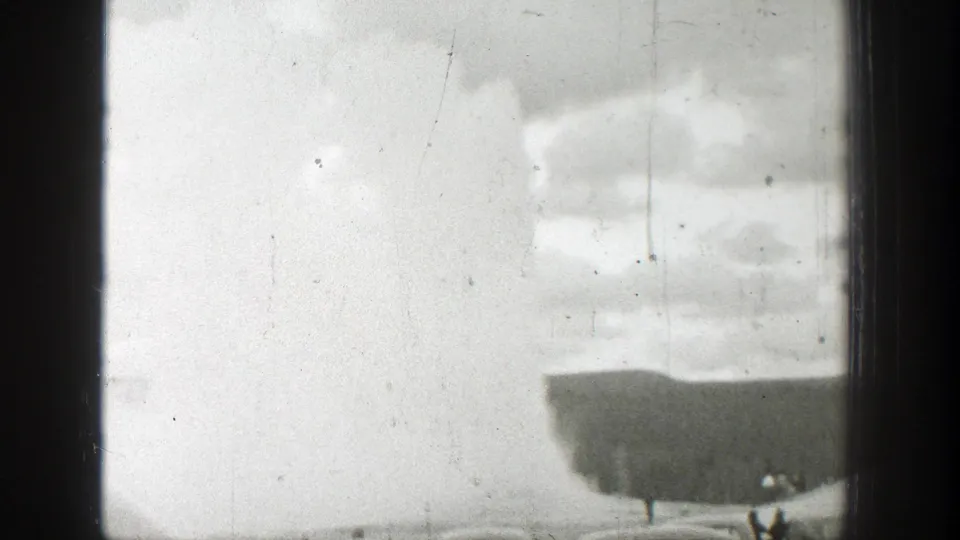 1939 Wyoming home-movie frame with a pale geyser plume, cloudy sky, low ridge, and small observers near the bottom edge.