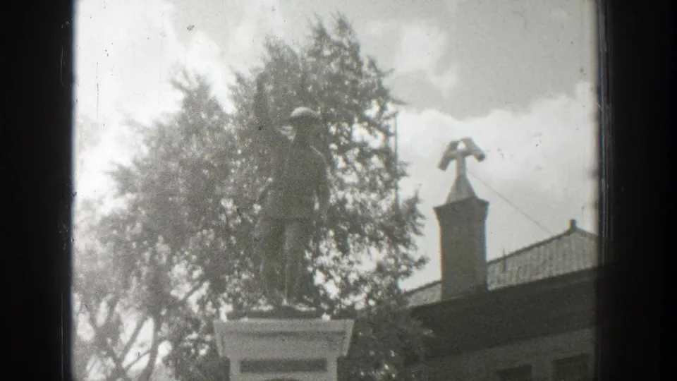 Black-and-white 1939 Wyoming home-movie frame showing a memorial statue on a pedestal, trees, building roofline, and cross-topped spire.