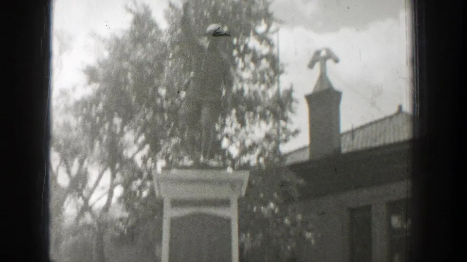 Black-and-white 1939 Wyoming frame showing the memorial statue more fully, with trees, a low building, railing, and cross-topped spire.