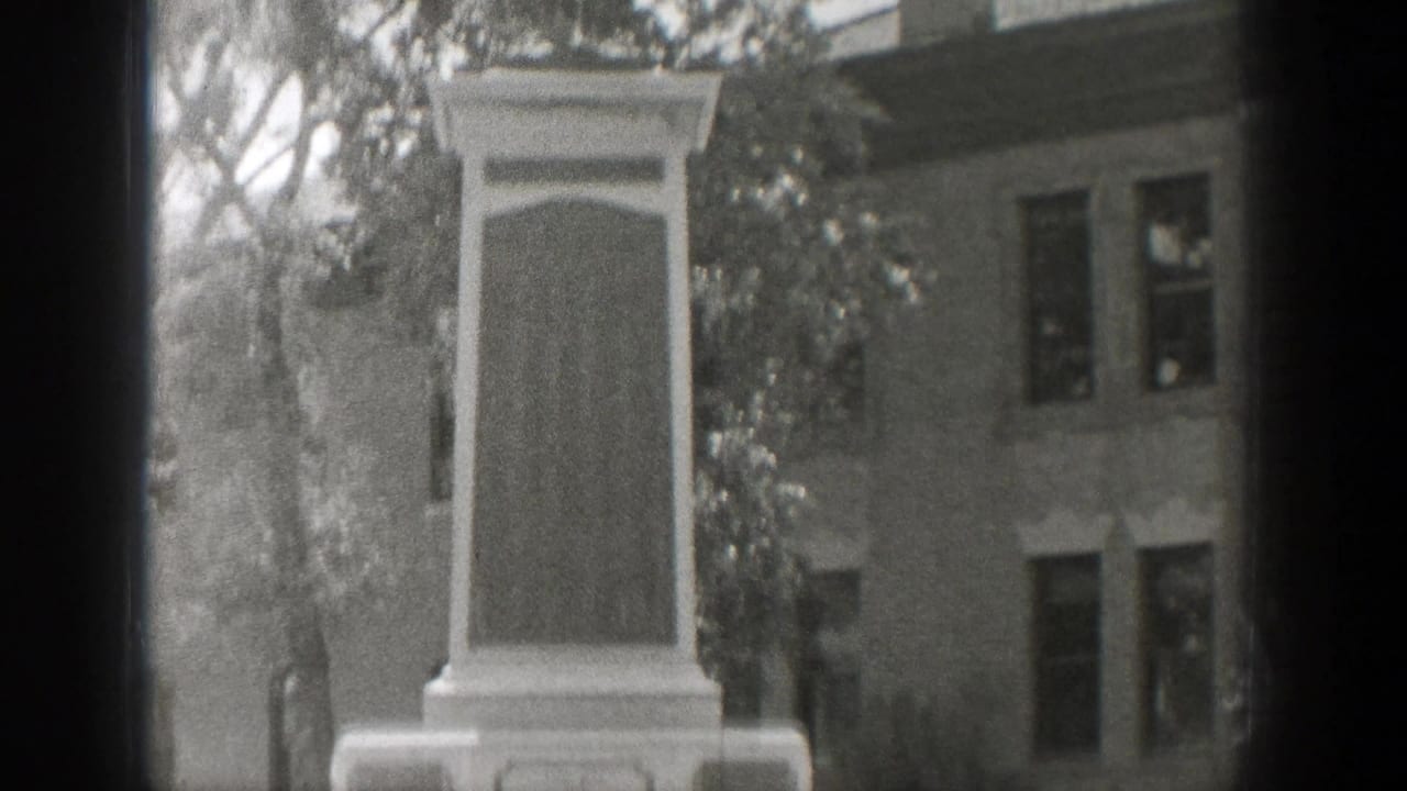 Stone monument centered beside trees with a building facade behind it.