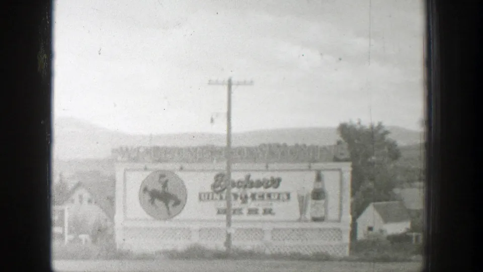 Black-and-white 1939 Wyoming home-movie frame of the Beckers Club Beer sign set against houses, trees, a utility pole, and hills.