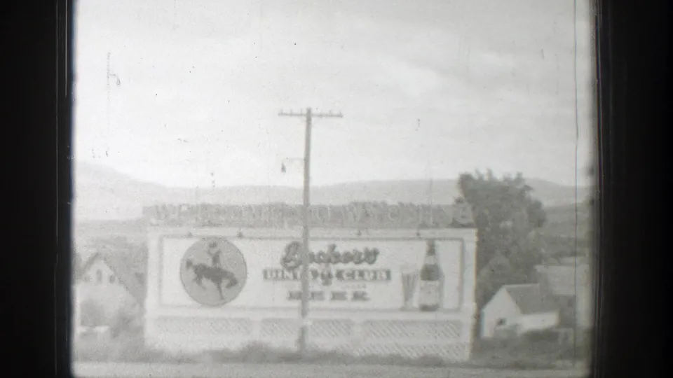 1939 Wyoming 8mm frame showing the billboard slightly lower in frame, with Beckers lettering, cowboy emblem, utility pole, houses, and hills.