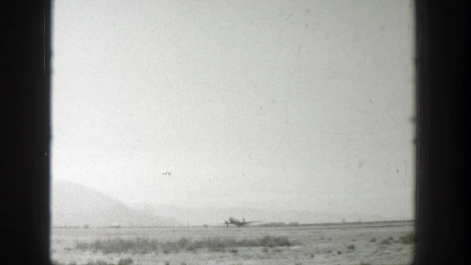 1939 Wyoming home-movie still of a distant propeller airplane on a grassy airfield, with low mountains in the background.