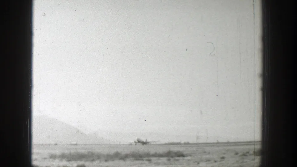 Black-and-white 8mm still from Wyoming in 1939, showing a small airplane low on the horizon across a rural airfield.