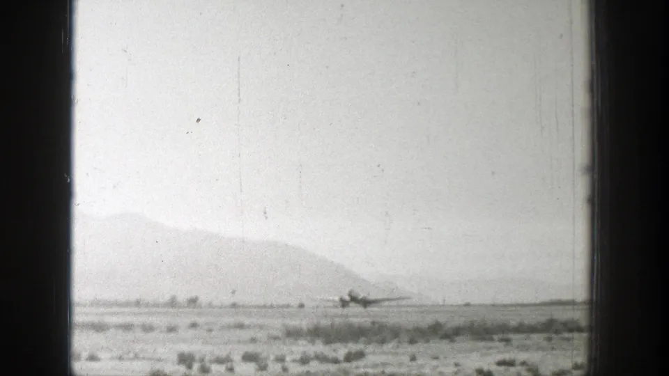 1939 Wyoming home-movie frame showing a distant propeller airplane on a rural airstrip below hazy mountains.