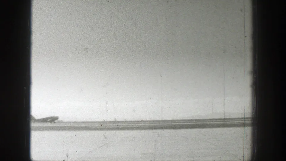Washed-out 8mm frame from 1939 Wyoming showing the airplane low and distant against the flat airfield.