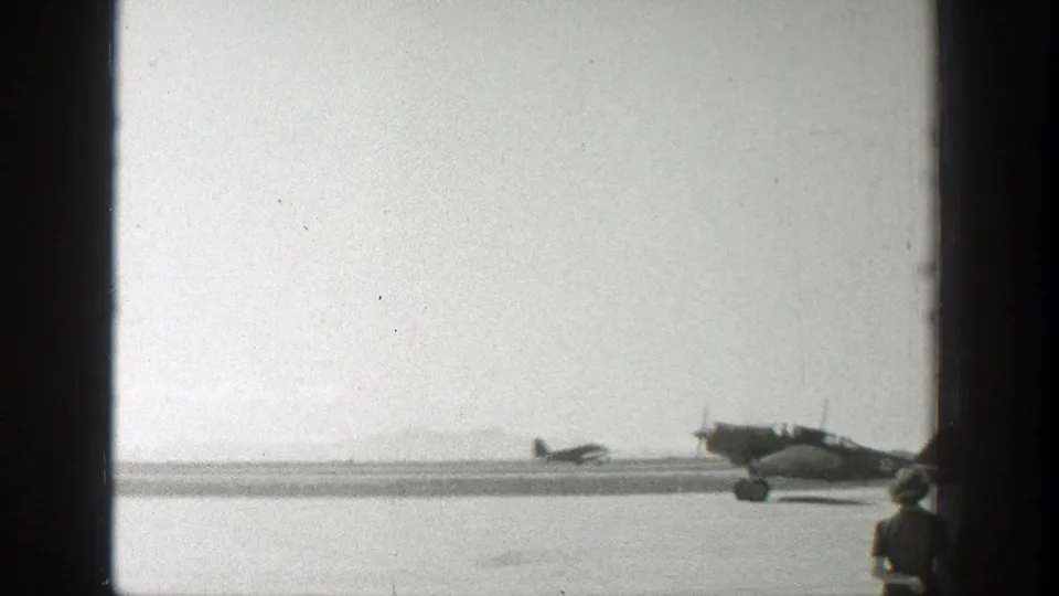 A foreground observer in a hat appears at the edge of a 1939 Wyoming 8mm airfield frame with propeller planes beyond.