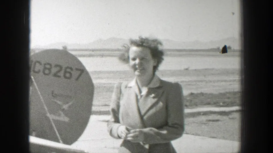 1939 Wyoming frame showing a woman in a tailored suit standing beside a marked aircraft tail near the runway.