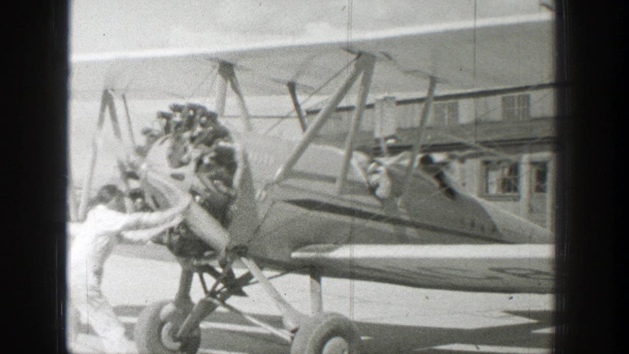 In supported 1939 Wyoming home-movie footage, a man in a light-colored jumpsuit stands by the radial engine and propeller of an open-cockpit biplane at an airfield.