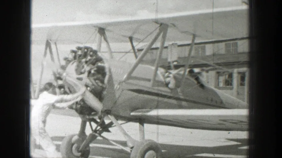 In supported 1939 Wyoming home-movie footage, a man in a light-colored jumpsuit stands by the radial engine and propeller of an open-cockpit biplane at an airfield.